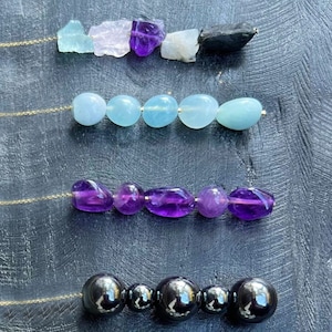 May include: A selection of gemstone and metal beads for a necklace. The beads include amethyst, aquamarine, rose quartz, moonstone, black onyx, clear quartz, jade, and gold hexagons. Add More Drops to Your Pull Me Necklace.