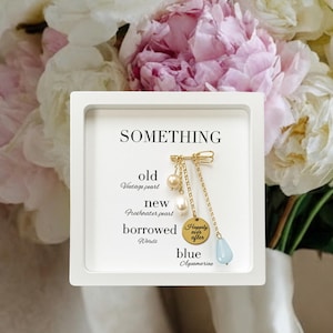 Puede incluir: Un recuerdo de boda enmarcado en blanco con la palabra «SOMETHING» en la parte superior. Debajo, el texto enumera «old, new, borrowed, blue». Un dije dorado con perlas y una piedra aguamarina azul cuelga del marco.