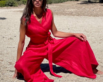 May include: A vibrant red sleeveless jumpsuit featuring a wrap-style bodice with a V-neckline and a matching fabric tie belt at the waist. The garment has wide-leg trousers that drape loosely, creating a flowing silhouette in a solid red color.