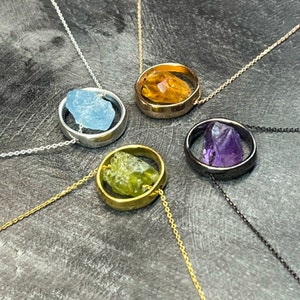 Peut inclure: Cinq colliers avec des pierres de différentes couleurs serties dans un pendentif circulaire. Les pierres sont de l'aigue-marine, de la citrine, du péridot, de l'améthyste et d'une pierre transparente. Les colliers sont sur un fond gris.
