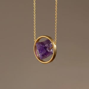 Pode incluir: Um colar de tom dourado com uma corrente fina e um pendente circular. Suspenso no centro do círculo dourado encontra-se uma pedra preciosa de ametista bruta, de cor violeta profunda, com uma superfície irregular, facetada e texturada.