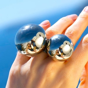 Könnte beinhalten: Zwei silberne Kugelringe werden auf einer Hand vor blauem Hintergrund gezeigt. Die Ringe spiegeln die Umgebung wider. Die größere Kugel befindet sich auf dem Zeigefinger, die kleinere auf dem Mittelfinger.