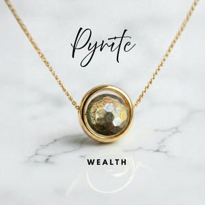 May include: A gold necklace with a pyrite gemstone pendant. The pyrite is set in a gold-tone circular frame. The word "Pyrite" is written in cursive above the pendant, and the word "WEALTH" is written below.