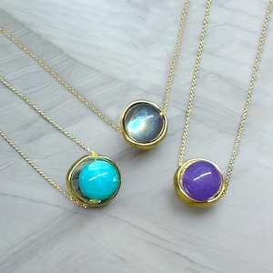 Può includere: Tre collane con sottili catenine dorate e pendenti circolari. Ogni pendente contiene una pietra preziosa liscia e rotonda: una grigia iridescente, una turchese brillante e una viola intenso. Le pietre sono incastonate in metallo dorato.