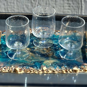 May include: Three clear, textured stemware glasses sit on a rectangular tray. The tray has a blue and gold resin surface with a gold-colored, textured border. The glasses are empty and have a unique, bubbly texture.