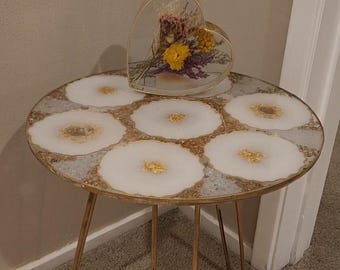 May include: A round side table with a gold metal frame and legs. The tabletop features a white and gold resin design with a heart-shaped glass container filled with dried flowers. The table is approximately 20 inches in diameter.