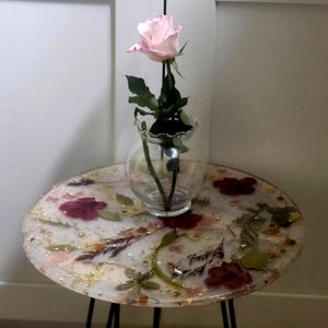 May include: A round side table with a clear resin top, embedded with dried flowers and gold flakes. A small glass vase holds a single pink rose. The table has black metal hairpin legs.