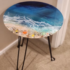 May include: Round side table with a resin top depicting an ocean scene. The top features blue and white waves, a sandy beach, and colorful shells. The table has black hairpin legs.