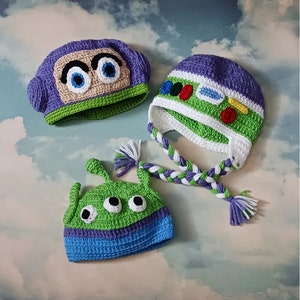 May include: Three handmade crochet hats in the style of characters from the animated film Toy Story. The hats include a purple Buzz Lightyear hat, a green and blue alien hat, and a Buzz Lightyear hat with a braided strap.