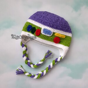 May include: A handmade crochet hat in the style of a cartoon character. The hat is primarily purple, green, and white, with colorful accents. Braided ties hang from the sides. The text "YGoCozyKnots" is visible on the side of the hat.