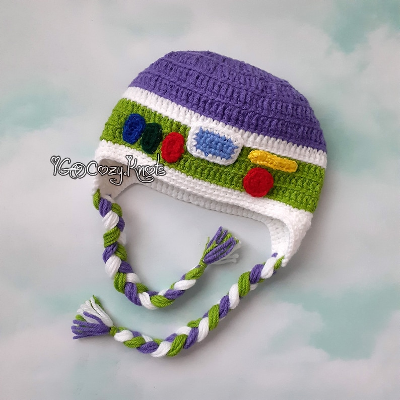May include: A handmade crochet hat in the style of a cartoon character. The hat is primarily purple, green, and white, with colorful accents. Braided ties hang from the sides. The text "YGoCozyKnots" is visible on the side of the hat.