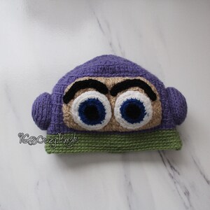 May include: A handmade crochet hat in the shape of a cartoon character's head. The hat is primarily purple, with large, expressive eyes in white, blue, and black. The base of the hat is green.