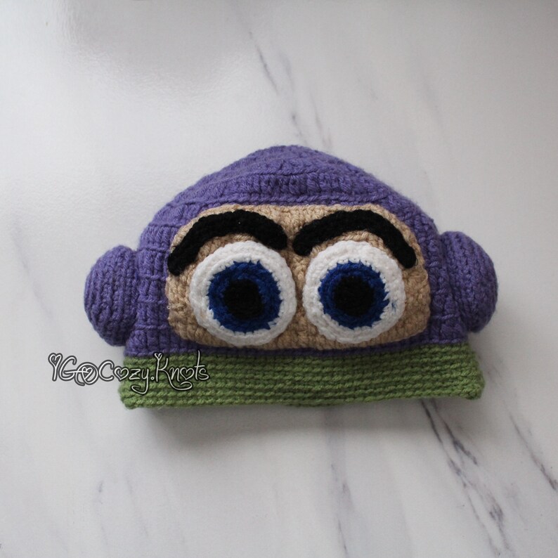 May include: A handmade crochet hat in the shape of a cartoon character's head. The hat is primarily purple, with large, expressive eyes in white, blue, and black. The base of the hat is green.