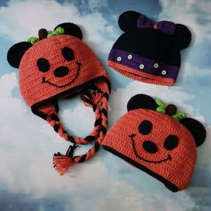 May include: Three handmade crochet hats. Two are orange with black accents, resembling pumpkins with smiling faces and mouse ears. The third is black with a purple band and bow, also with mouse ears. The hats are likely for Halloween or themed events.