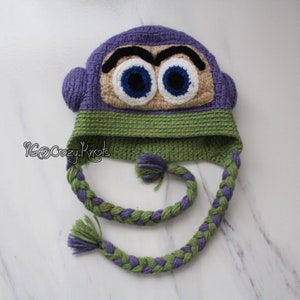 May include: A handmade crochet hat in the shape of a cartoon character's face. The hat features a purple top, green band, and braided ear flaps. The character's face has large blue eyes, black eyebrows, and a tan face.