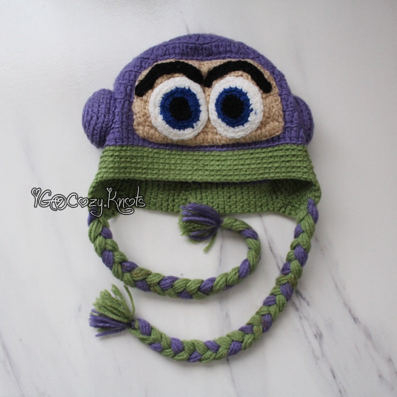 May include: A handmade crochet hat in the shape of a cartoon character's face. The hat features a purple top, green band, and braided ear flaps. The character's face has large blue eyes, black eyebrows, and a tan face.