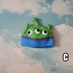 May include: A handmade crochet hat in the shape of a green alien with three eyes. The hat features a blue brim with a purple stripe and three white and black eyes. The hat has two antennae and a single antenna on top.