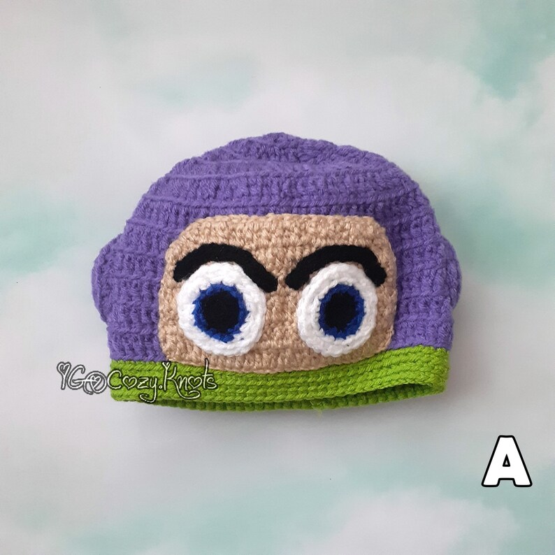 May include: A crocheted hat in the style of a cartoon character. The hat is primarily purple with a tan face, green trim, and large blue and white eyes. The hat is handmade and features a detailed design.