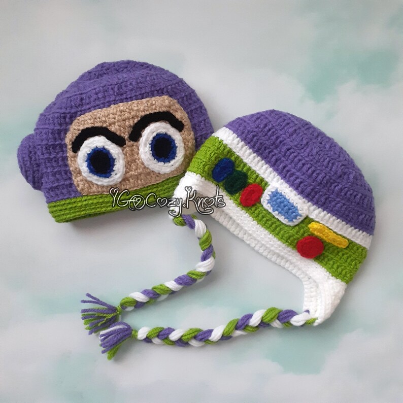 May include: Two handmade crochet hats. One is purple with a tan face and large blue eyes, resembling a character. The other is purple, green, and white, with colorful accents and a braided tie. The hats are designed to look like a character.