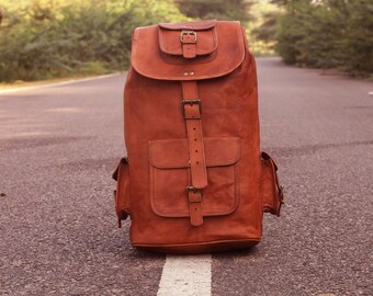May include: A brown leather backpack with multiple pockets and buckles. The backpack is sitting on a paved road.