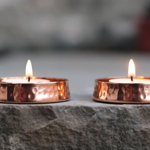 May include: Two round tea light candle holders made of polished copper with a hammered texture. Each holder contains a small white tea light candle with a lit flame, casting a warm glow on the metallic surface of the copper containers.