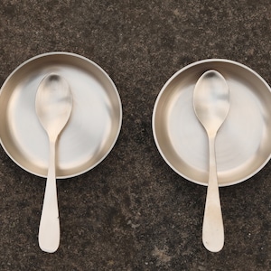 May include: Two silver spoons are placed inside two silver bowls. The bowls are sitting on a grey textured surface.
