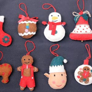 Puede incluir: Ocho adornos navideños de fieltro con varios diseños, incluyendo una media, un hombre de jengibre, un muñeco de nieve, un elfo, una campana y un oso. Los adornos son todos rojos, verdes, marrones y blancos.