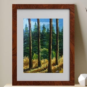 May include: A framed landscape painting depicts a forest scene with tall, slender trees and a backdrop of green foliage and a blue sky. The artwork is encased in a dark brown wooden frame and rests on a light-colored shelf.