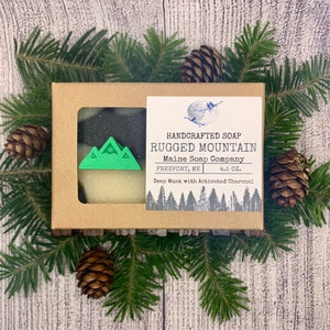 May include: A brown cardboard box with a clear window showing a bar of soap with black and white swirls. The box has a white label with the text "HANDCRAFTED SOAP RUGGED MOUNTAIN Maine Soap Company FREEPORT, ME 4.5 OZ. Deep Musk with Activated Charcoal".