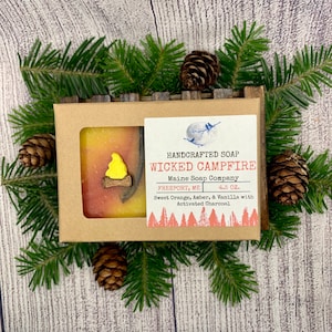 May include: A brown cardboard box with a bar of soap inside. The soap is orange and yellow with a wooden campfire design. The box has a label that reads "HANDCRAFTED SOAP WICKED CAMPFIRE Maine Soap Company FREEPORT, ME 4.5 oz. Sweet Orange, Amber, & Vanilla with Activated Charcoal".