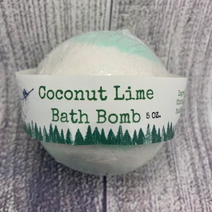 May include: A white bath bomb with a light green top, wrapped in clear plastic. The bath bomb is labeled "Coconut Lime Bath Bomb 5 oz." with a green forest design along the bottom of the label. The background is a light wood grain.