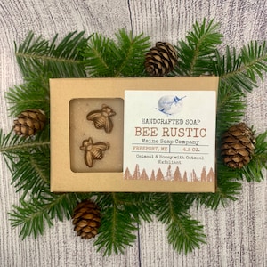May include: A brown cardboard box with a clear plastic window showing a bar of soap with two gold bee-shaped embellishments. The box has a label that reads "HANDCRAFTED SOAP BEE RUSTIC Maine Soap Company FREEPORT, ME 4.5 OZ. Oatmeal & Honey with Oatmeal Exfoliant".