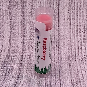 May include: A pink lip balm tube with a white label that reads "Raspberry Maine Soap Company". The label has a green illustration of pine trees.