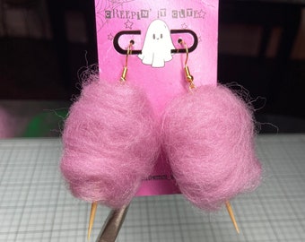 May include: Pink cotton candy earrings with gold-colored hooks. The earrings are attached to a pink card with the text "CREEPIN' IT CUTE" and a ghost illustration. The earrings are held by a pair of pliers.