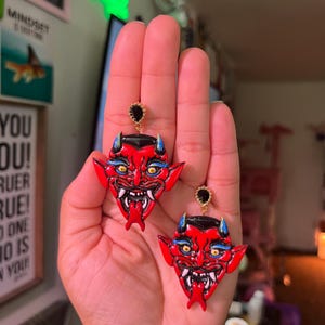 May include: A pair of red devil face earrings with black eyes, blue eyebrows, and a gold accent. The earrings are hanging from a person's hand.