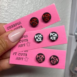 May include: Three pairs of round stud earrings, each black with a contrasting red or white design. The earrings are presented on pink cards, with the text "CREEPIN CUTE ART" printed on them.