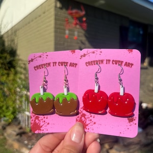 May include: Two pairs of dangle earrings, each with a candy apple design. One pair features brown and green apples, the other red. The earrings are displayed on pink cards with the text "CREEPIN' IT CUTE ART".