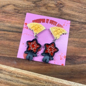 May include: A pair of dangle earrings featuring a waffle design with a bite taken out of it, connected to a red star-shaped charm with white dots and a gray figure. The earrings are displayed on a pink card with the text "CREEPIN IT CUTE ART".