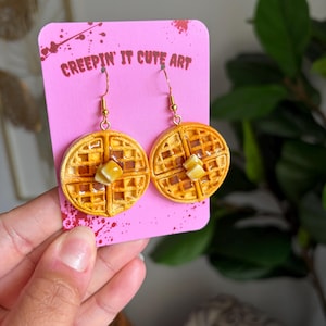 May include: A pair of dangle earrings shaped like round waffles with a golden-brown texture, topped with a small square of butter and a glossy syrup finish. The earrings are attached to a bright pink display card with the text CREEPIN' IT CUTE ART.