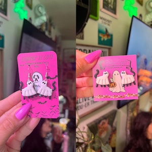 Op de afbeelding: Roze kaart met handgemaakte draagbare kunst. De kaart toont witte spookoorbellen en -spelden met zwarte kattenstaarten. De tekst "CREEPIN' IT CUTE" is zichtbaar. De oorbellen en spelden zijn Halloween-thema.