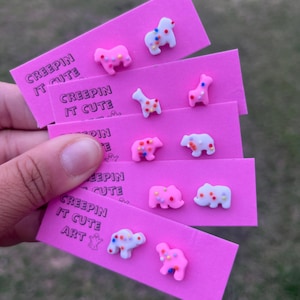 Peut inclure: Cartes de boucles d'oreilles roses avec le texte "CREEPIN IT CUTE ART" et des boucles d'oreilles clous en forme d'animaux. Les boucles d'oreilles sont blanches et roses avec des pois colorés.