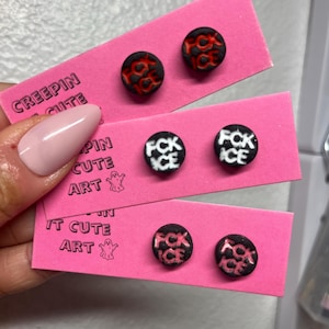 May include: Three pairs of round stud earrings displayed on pink cards. The earrings have black and red, black and white, and black and pink designs with the text "FCK ICE" or a similar design. The cards are printed with the text "CREEPIN CUTE ART".