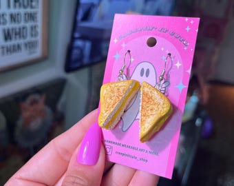 May include: A pair of grilled cheese sandwich earrings on a pink card with the text "handmade wearable art & more. creepinitcute_shop".