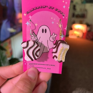 Pode incluir: Um par de brincos feitos à mão com um bolo rosa e branco com cobertura de chocolate. Os brincos estão em um cartão rosa com o texto "Creepin' It Cute" e uma ilustração de um fantasma. O cartão também inclui o texto "Handmade Wearable Art & More." e "creepinitcute_shop".