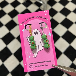 Op de afbeelding: Een paar groene olijven oorbellen met gouden en zilveren haken. De oorbellen zijn op een roze kaart met de tekst "Creepin' It Cute" en "Handmade Wearable Art & More." met de Instagram-handle "creepinitcute_shop".
