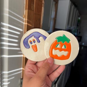 Könnte beinhalten: Zwei runde Halloween-Untersetzer. Einer zeigt einen lila Geist mit orangefarbenem Mund und Augen, der andere einen orangefarbenen Kürbis mit grünem Stiel. Beide haben einen cremefarbenen Hintergrund.