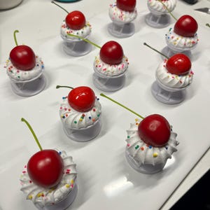 Puede incluir: Un conjunto de agarres para teléfono con cerezas. Cada agarre presenta una cereza roja brillante sobre un remolino de crema batida blanca, decorada con chispas de colores. Los agarres son blancos y tienen un diseño plegable.