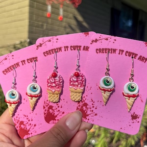 Puede incluir: Pendientes rosas con temática espeluznante. Los pendientes presentan un cerebro, un globo ocular y un diseño de cono de helado. Los pendientes están en tarjetas rosas con el texto "CREEPIN' IT CUTE ART".