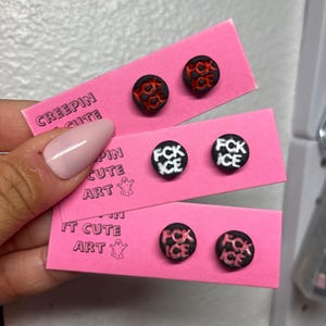 May include: Three pairs of round stud earrings on pink cards. The earrings have black backgrounds with the words "FCK ICE" in white or red. The cards have the text "CREEPIN CUTE ART".