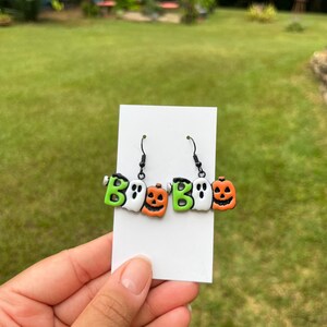 May include: Halloween-themed earrings featuring the word "Boo" with a ghost and pumpkin design. The earrings have black hooks and are displayed on a white card. The word "Boo" is in green and white, and the pumpkins are orange.